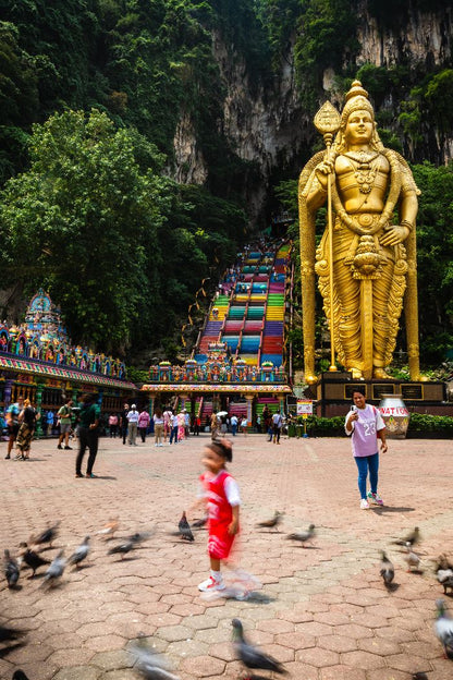 Child dancing near pigeons in front of the golden Murugan statue and colorful staircase at Batu Caves, Kuala Lumpur, Malaysia